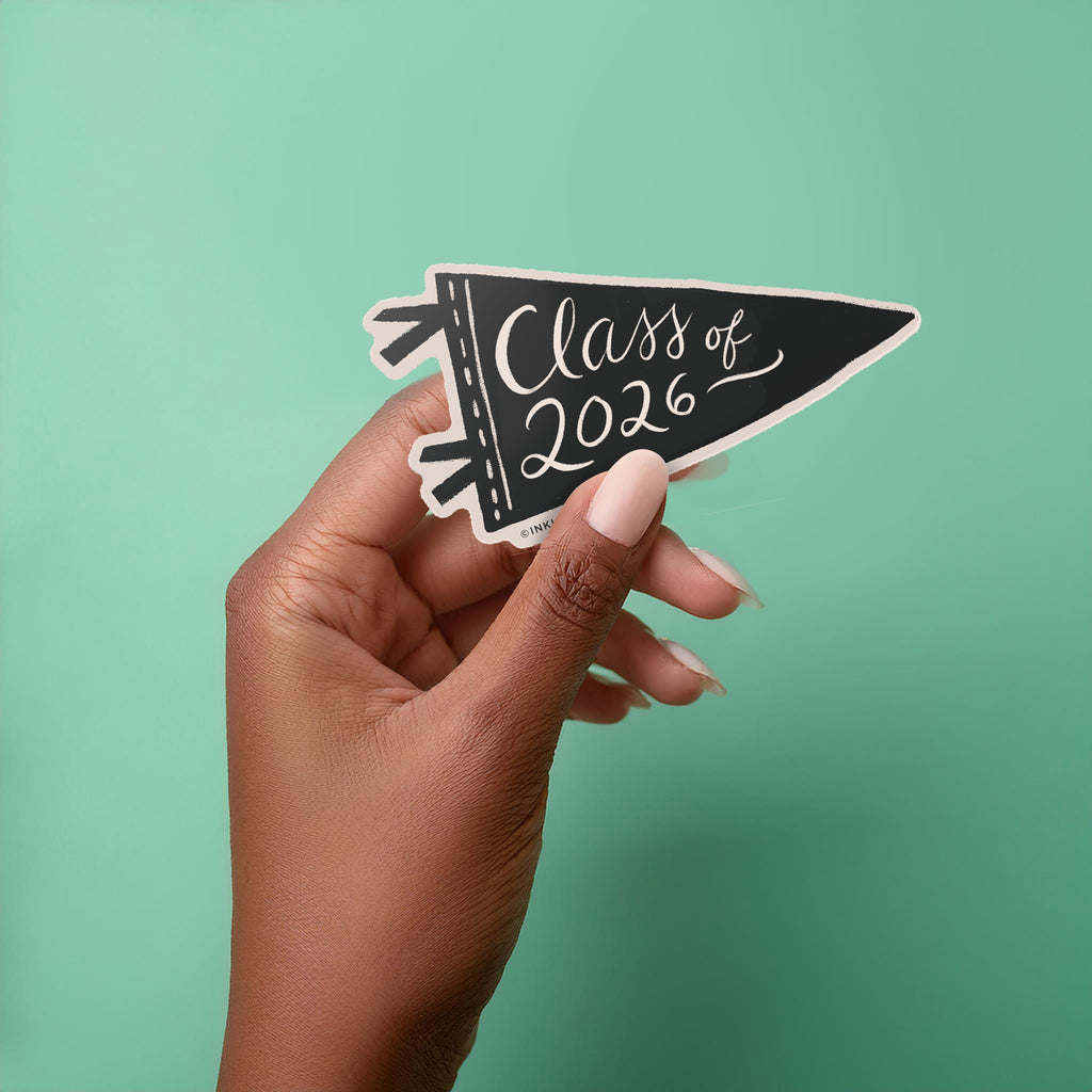 Hand holding a vinyl sticker in a black pennant shape with "Class of 2026" in a hand-lettered script text on a white background