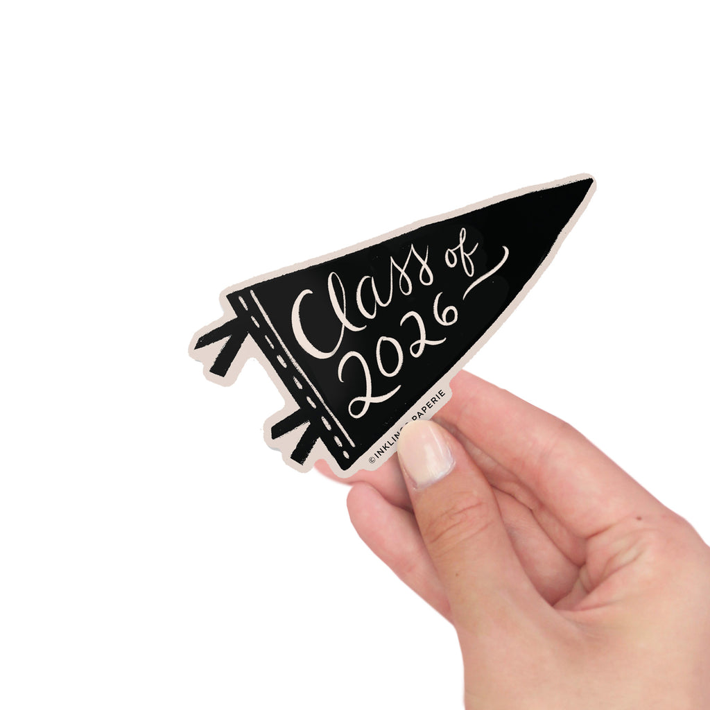 Hand holding a Class of 2026 vinyl sticker in a black pennant shape with script text on awhite background