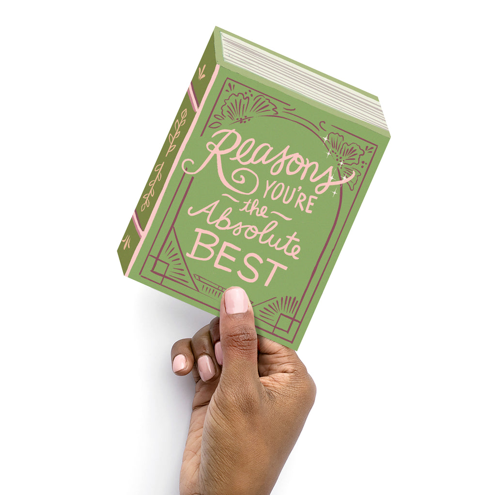 Hand holding a book-shaped greeting card with die cut corners and hand-lettered message on the front that reads, "Reasons You're the Absolute Best"